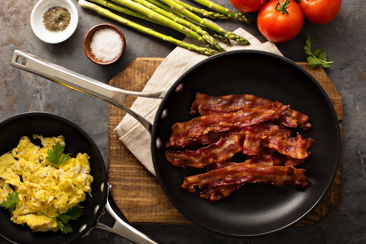 A pan of crisp bacon alongside other breakfast items, including eggs, tomatoes, and asparagus.