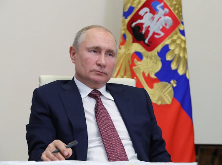 Vladimir Putin sits with pen in his hand in front of a Russian Federation flag.