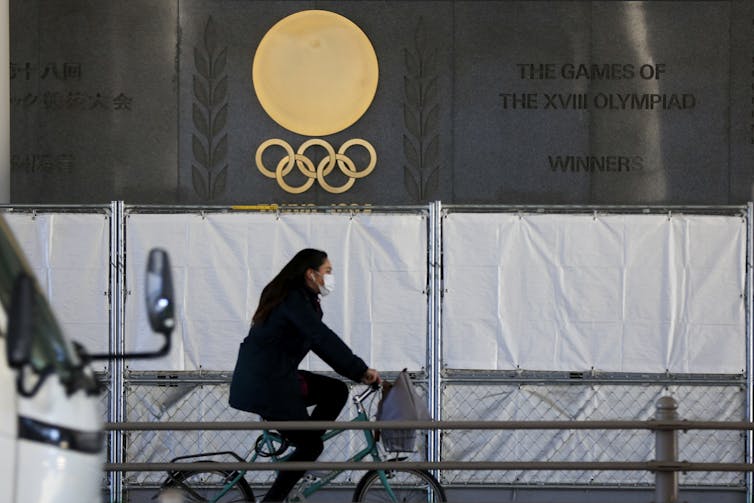 A woman wearing a face mask riding a bicycle past an Olympics logo