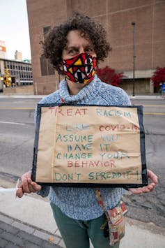 Una mujer con una máscara sostiene un cartel que compara el COVID-19 con el racismo: 'suponga que lo tiene'