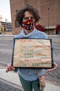 Woman wearing a mask holds a sign likening COVID-19 to racism – 'assume you have it'