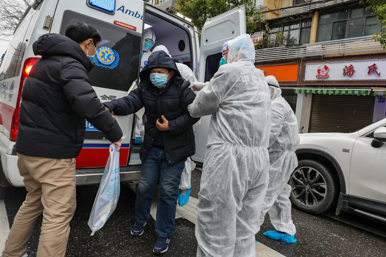Medical staff in full body protective suits help man with face mask out of ambulance.