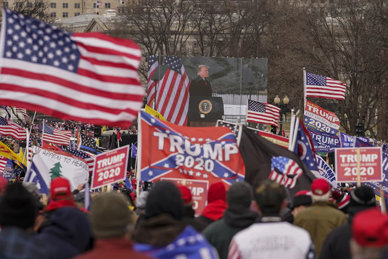 Trump stands behind a clear partition, surrounded by supporters waving assorted flags.