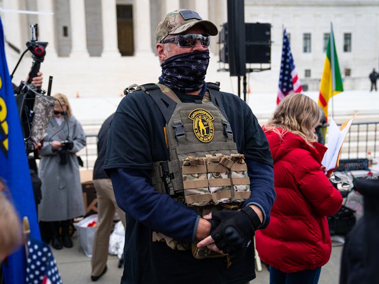 Man in camouflage, a bulletproof vest and sunglasses stands guard with hands folded