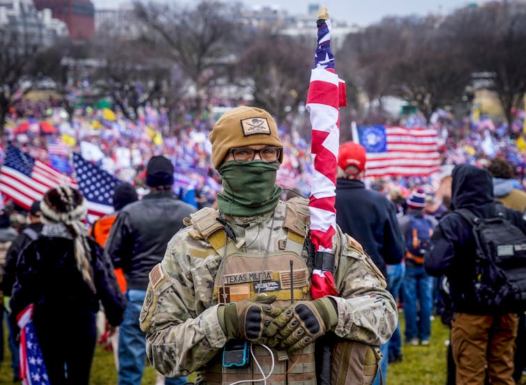 Glasses-wearing man in military fatigues poses with an American flag in front of a large crowd