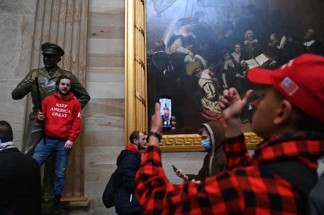 A man uses a cell phone to take a photo of another man standing on the pedestal of a statue in the U.S. Capitol building.