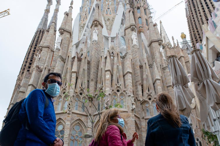 Three people wearing masks standing in front of La Sagrada Familia cathedral in Barcelona, Spain.