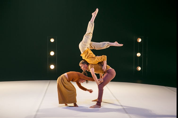 Two circus performers stand stooped over; a third lies on their backs with her feet in the air.