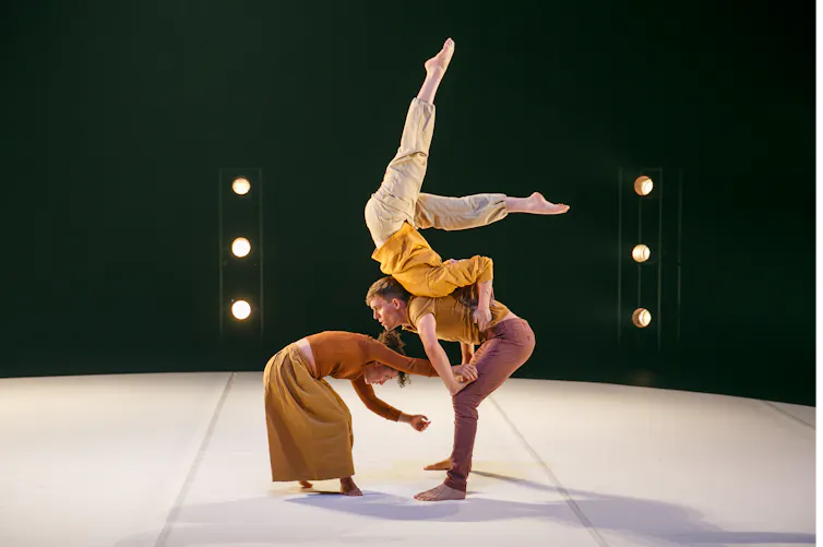 Two circus performers stand stooped over; a third lies on their backs with her feet in the air.