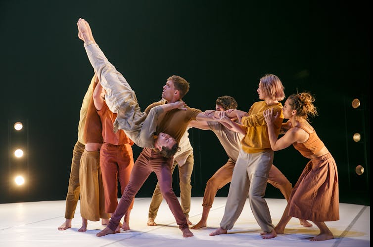 Eight people stand right-way up, holding one man upside down.