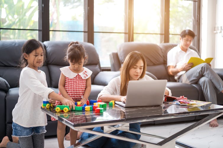 Family scene with two daughters playing, mother working on laptop, father reading