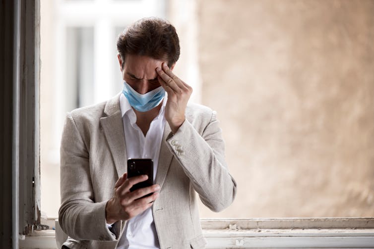 Man in medical face mask holds head and looks at phone in confusion