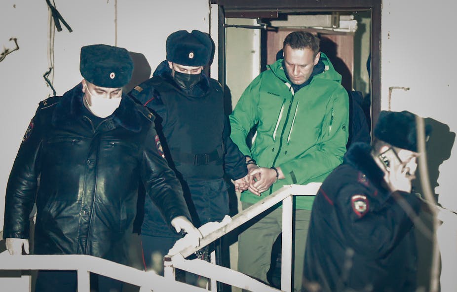 Russian police officers in masks surround Alexei Navalny, wearing a green jumpsuit and manacles