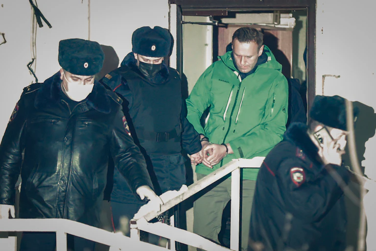 Russian police officers in masks surround Alexei Navalny, wearing a green jumpsuit and manacles