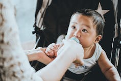 A baby looks at his mother while sucking on his bottle in a stroller.