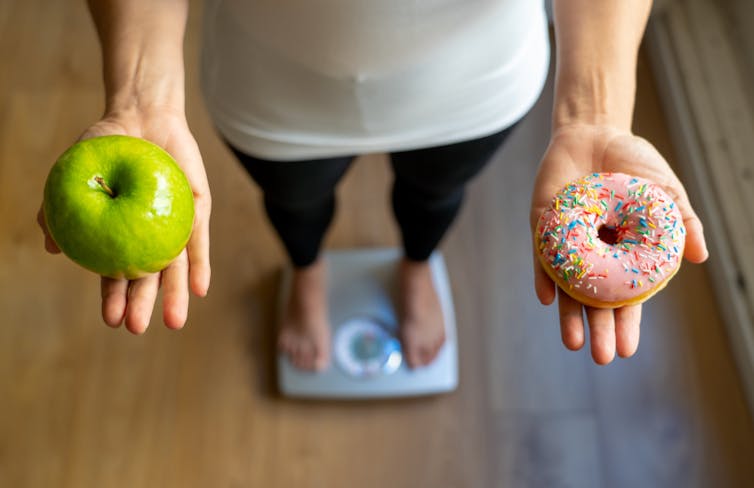 A person stands on the scales, holding an apple in one hand, and a donut in the other.