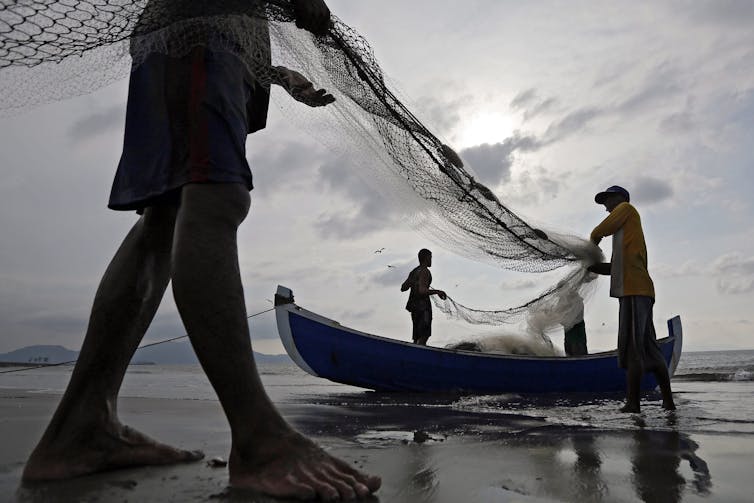 Three men haul fishing nets by a small boat at a beach.