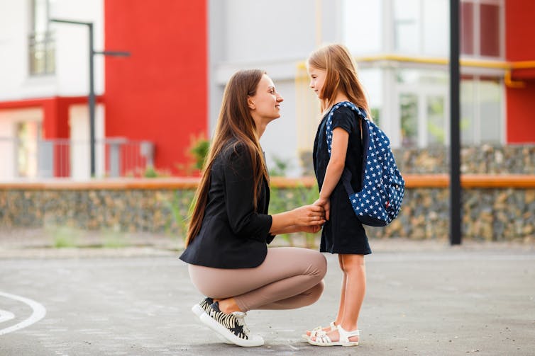 Outdoors, a woman kneels by her young daughter wearing a backpack.