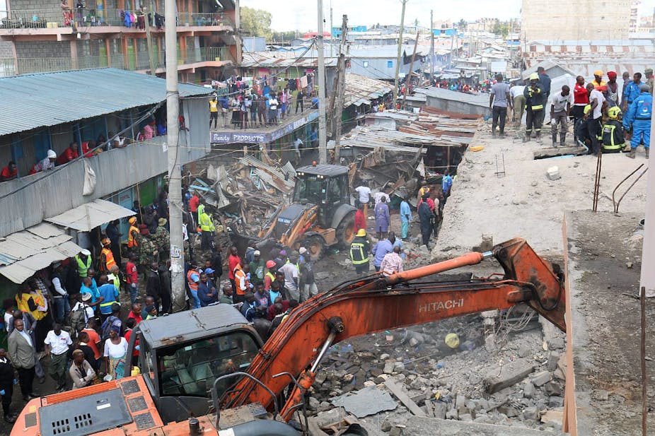 Groups of people gather around building rubble while a huge orange digger lifts debris