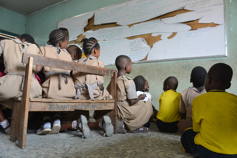 Children sitting in a classroom and facing a shabby white board.
