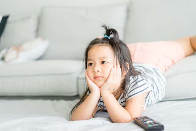 A young girl watches television with a remote on the ground next to her