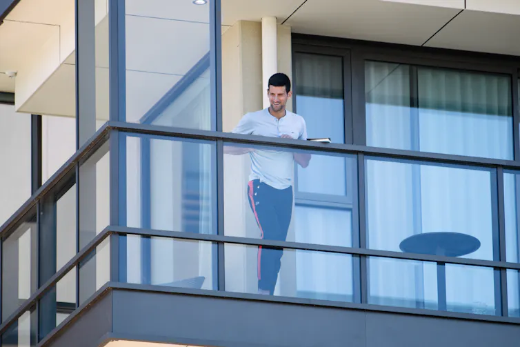 Tennis player Novak Djokovic leans on a glass balcony.