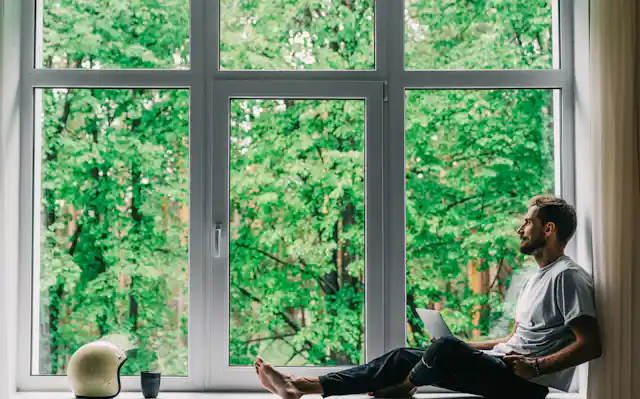 A man site in a window with a laptop on his legs.