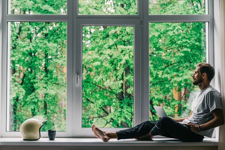 A man site in a window with a laptop on his legs.