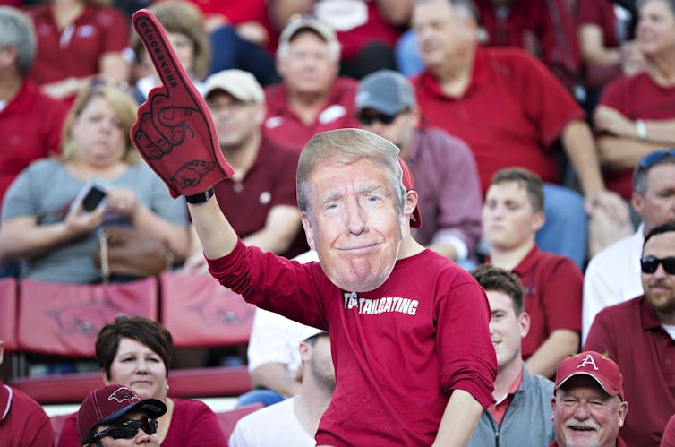 man at sporting event wearing a trump face mask and holding rubber finger.