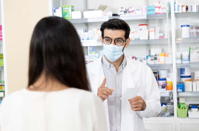 Male pharmacist wearing mask hold prescription speaking to customer