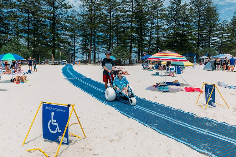 A blue mat cuts across the white sandy beach. A woman smiles in a beach wheelchair.