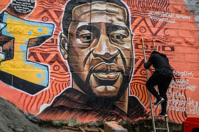 A mural artist climbs up a ladder leaning against a wall that features a mural of a black man, eyes cast downward.