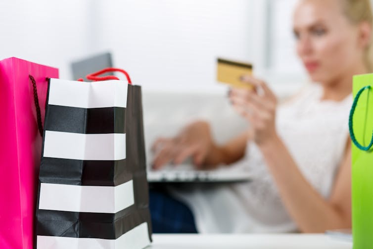 Blond woman making purchasing via internet paying credit card. Focus on fresh buying packed in colored paper bags standing in front of her. Shopping, consumerism, delivery and present concept