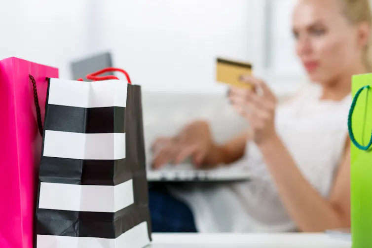 Blond woman making purchasing via internet paying credit card. Focus on fresh buying packed in colored paper bags standing in front of her. Shopping, consumerism, delivery and present concept