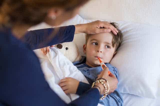 Una mujer pone un termómetro en la boca a un niño que está en la cama con su oso de peluche.