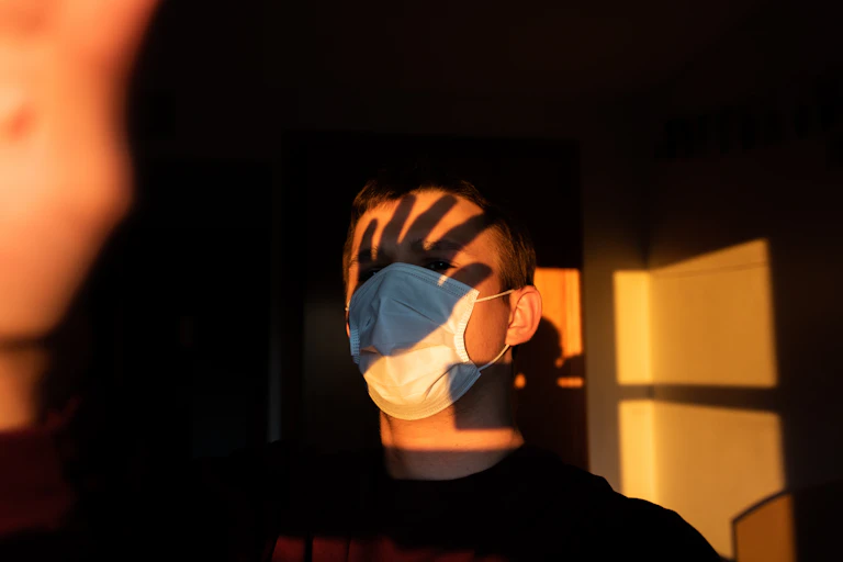 Sombra de una mano al caer la tarde sobre la frente de un hombre con mascarilla.