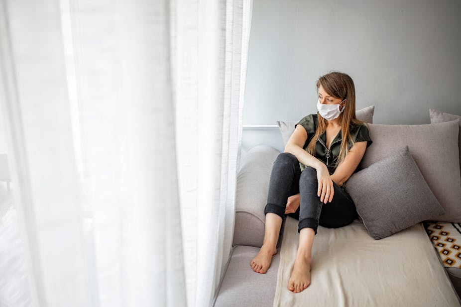 Woman wearing surgical mask sits in her home looking out the window.