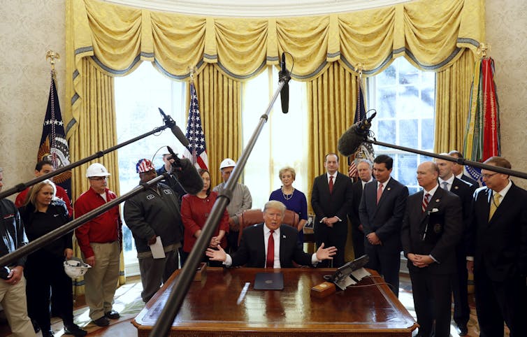 Trump is surrounded by manufacturers and media cameras in the Oval Office.