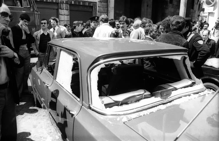 Black-and-white image of a crowd looking at a bullet-riddled car with shatters windshields and windows