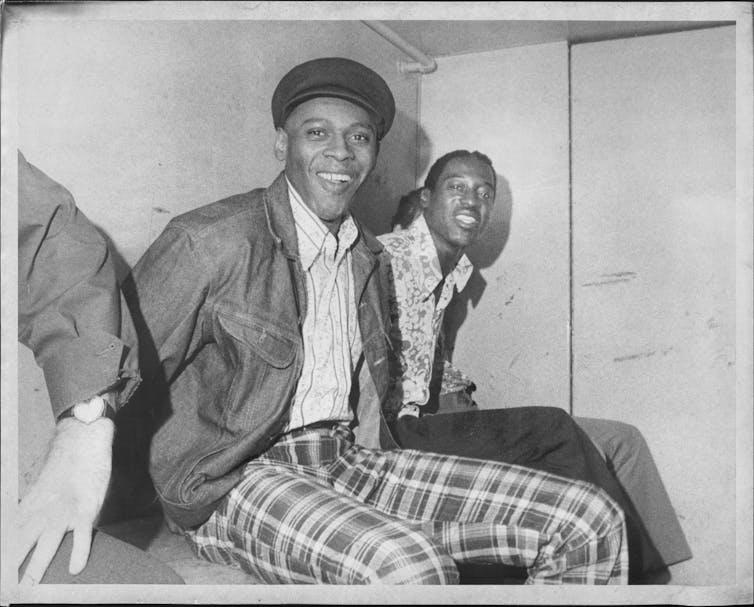 Two Black men in handcuffs in a paddy wagon smile at the camera
