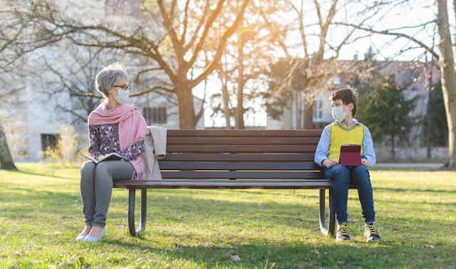 A woman and her grandson wearing face masks sitting on opposite ends of a park bench