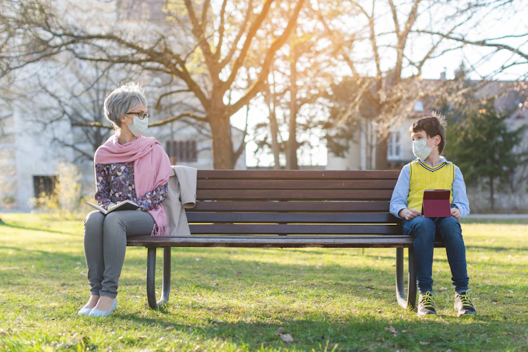 A woman and her grandson wearing face masks sitting on opposite ends of a park bench