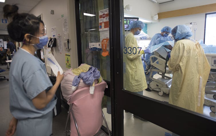 A health-care worker looking into an ICU room where other health-care workers are treating a patient