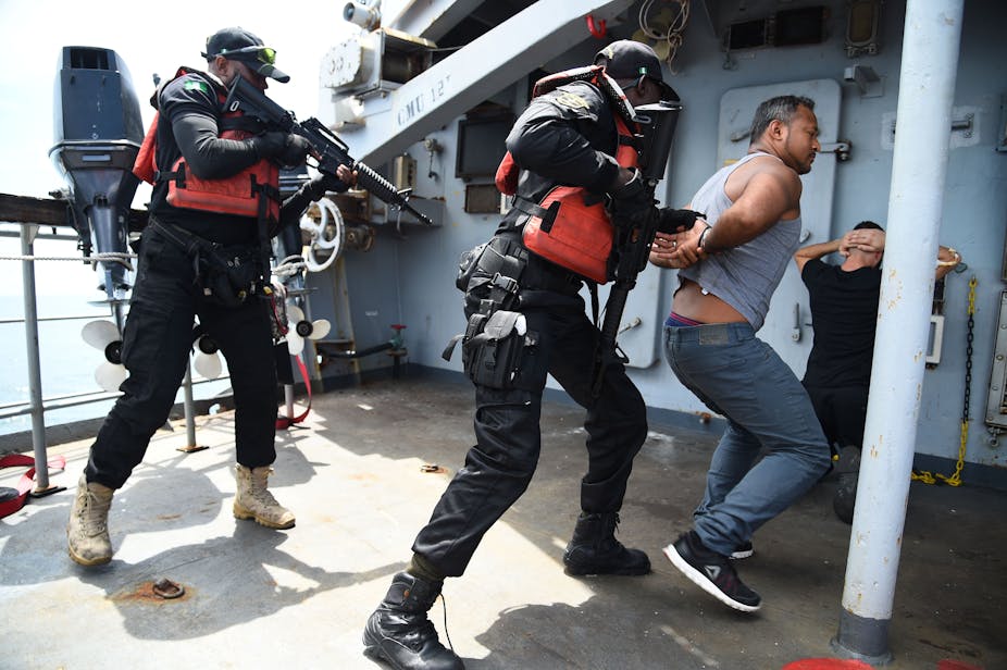 Men in black Nigerian Navy Special forces uniform point guns at two men pretending to be pirates in a staged arrest aboard a ship. 