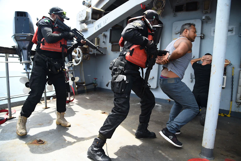 Men in black Nigerian Navy Special forces uniform point guns at two men pretending to be pirates in a staged arrest aboard a ship.