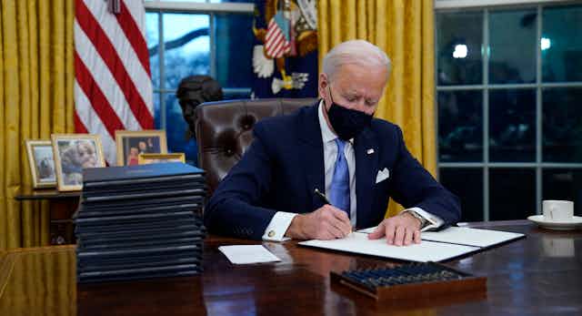 Biden, wearing a mask, signs executive orders in the Oval Office.
