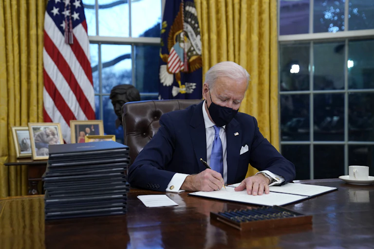 Biden, wearing a mask, signs executive orders in the Oval Office.