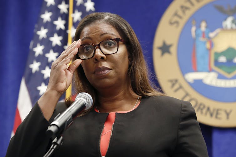 Photo of a woman wearing glasses, standing in front of a state crest and an American flag