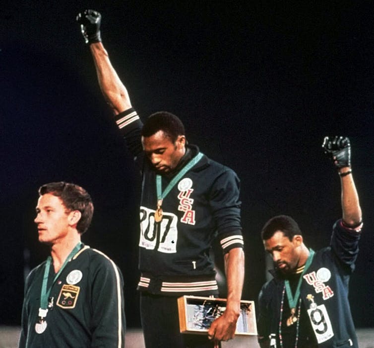 Australian silver medalist Peter Norman stands on the medal podium as Americans Tommie Smith and John Carlos raise their gloved fists in a human rights protest.