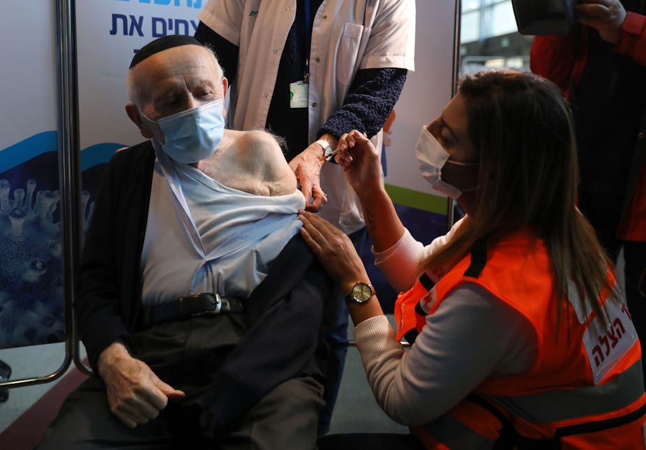 An elderly man wearing a yarmulke receives a vaccine from a young woman in a high-biz vest and mask.